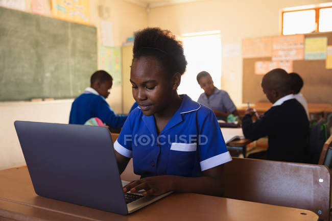 Vista frontal de perto de uma jovem estudante africana sentada em uma mesa usando um computador portátil e sorrindo durante uma aula em uma sala de aula da escola primária da cidade, nos colegas de classe de fundo estão sentados em suas mesas trabalhando — Fotografia de Stock