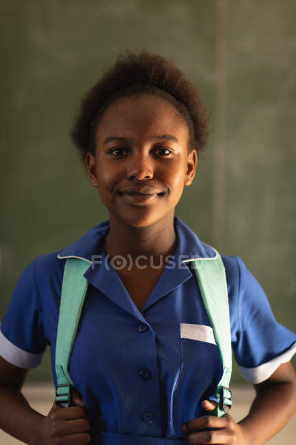 Ritratto da vicino di una giovane studentessa africana che indossa l'uniforme scolastica e lo zaino, guardando dritto verso la telecamera sorridente, in una scuola elementare cittadina — Foto stock