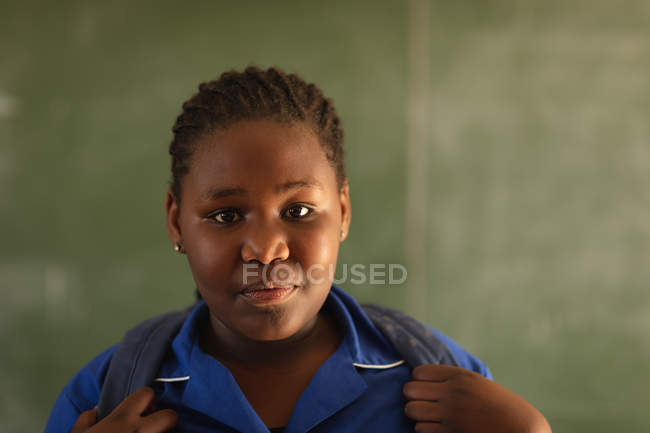 Retrato de perto de uma jovem estudante africana vestindo seu uniforme escolar e saco escolar, olhando diretamente para a câmera sorrindo, em uma escola primária da cidade — Fotografia de Stock