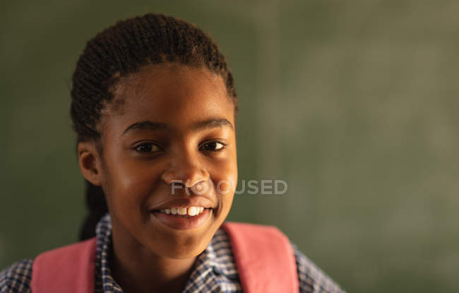 Portrait close up of a young African schoolgirl wearing her school uniform and schoolbag, looking straight to camera smiling, at a township elementary school — Stock Photo