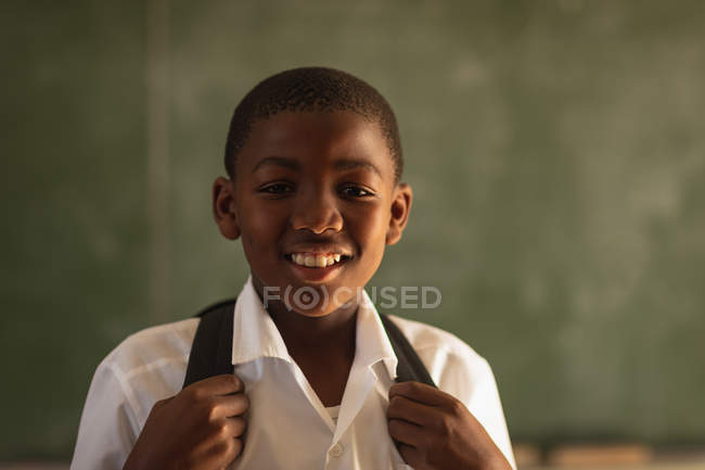 Портрет крупным планом молодого африканского школьника в школьной форме и школьной сумке, смотрящего прямо в камеру улыбающегося, на городскую начальную школу — стоковое фото