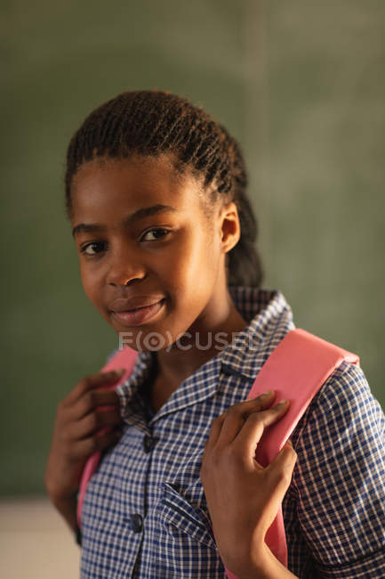 Retrato de perto de uma jovem estudante africana vestindo seu uniforme escolar e saco escolar, olhando diretamente para a câmera sorrindo, em uma escola primária da cidade — Fotografia de Stock