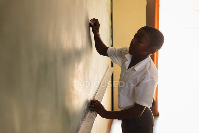 Vista lateral de perto de um jovem estudante africano em pé na frente da aula escrevendo no quadro negro durante uma aula em uma sala de aula da escola primária da cidade — Fotografia de Stock