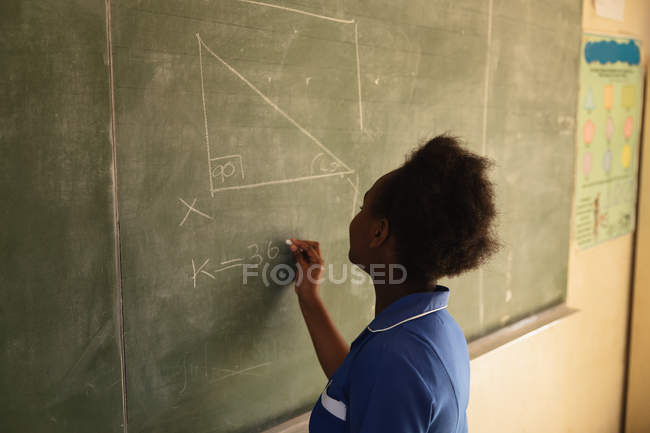 Вид сбоку на молодую африканскую школьницу, стоящую перед классом и пишущую на доске во время урока в городском классе начальной школы — стоковое фото