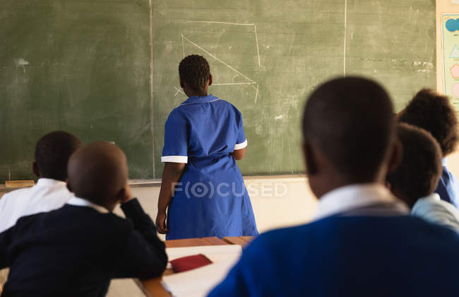Back view of a young African schoolgirl standing at the front of the class writing on the blackboard during a lesson in a township elementary school classroom. — Stock Photo