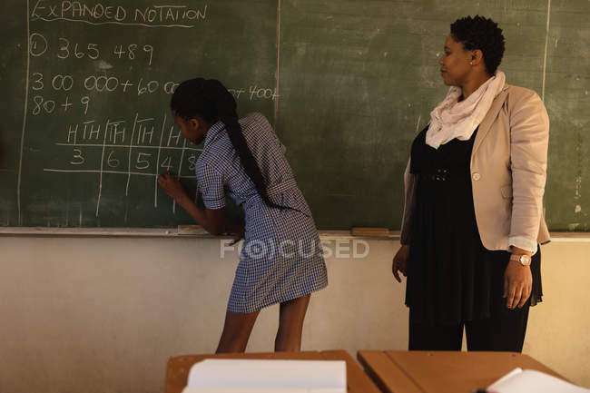 Back view of a young African schoolgirl standing at the front of the class writing on the blackboard with her middle aged African female teacher standing beside her watching, during a lesson in a township elementary school classroom — Stock Photo