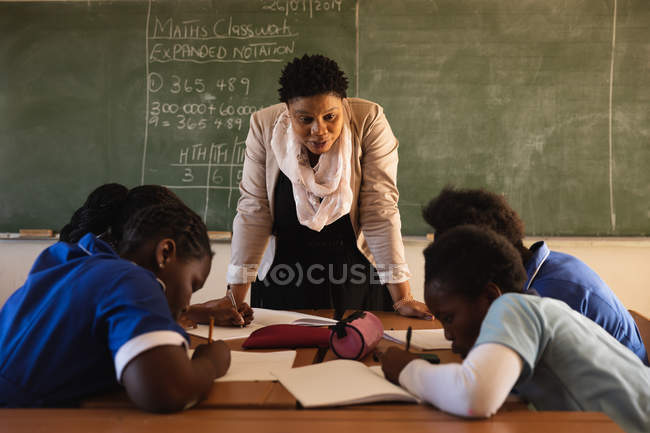 Front view close up of a middle aged African female school teacher standing at the front of the class in front of the blackboard and leaning forward to watch her pupils writing at their desks during a lesson in a township elementary school classroom — Stock Photo