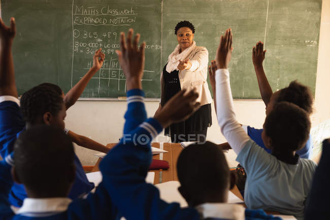 Front view of a middle aged African female school teacher standing at the front of the class in front of the blackboard with the rear view of pupils sitting at their desks with hands raised in the foreground during a lesson — Stock Photo