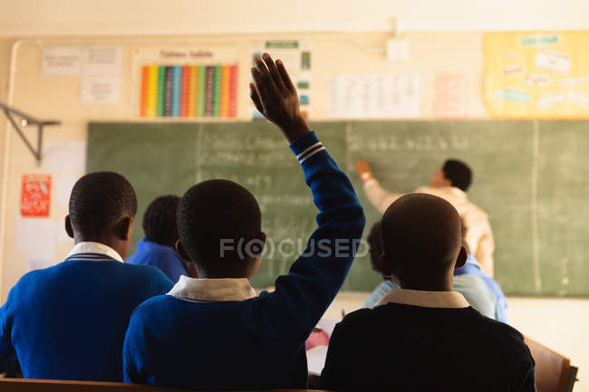 Rear view of pupils sitting at their desks with hands raised during a lesson in a township elementary school classroom with their teacher standing writing on the blackboard in the background — Stock Photo