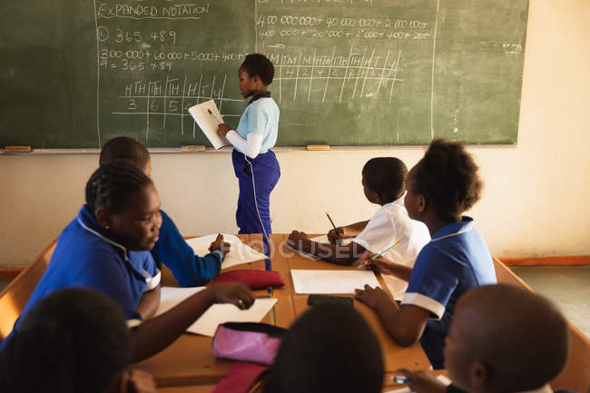 Vue latérale d'une jeune écolière africaine debout à l'avant de la classe tenant un livre et écrivant sur le tableau noir lors d'une leçon dans une classe de l'école élémentaire d'un canton . — Photo de stock