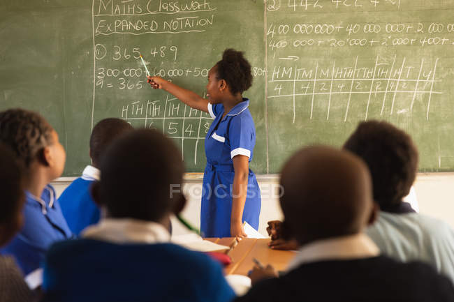 Side view of a young African schoolgirl standing at the front of the class writing on the blackboard during a lesson in a township elementary school classroom. — Stock Photo