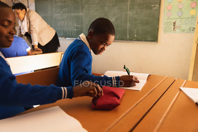 Vue latérale de deux jeunes écoliers africains assis à un bureau travaillant pendant une leçon dans une classe de l'école élémentaire d'un canton, en arrière-plan l'enseignant aide certains camarades de classe à leur bureau — Photo de stock