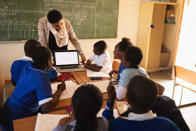 Front view of a middle aged African school teacher standing in front of the blackboard showing her pupils a laptop during a lesson in a township elementary school classroom. — Stock Photo