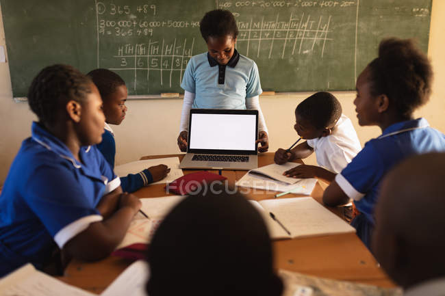 Front view of a young African schoolboy standing in front of the blackboard showing his classmates a laptop during a lesson in a township elementary school classroom. — Stock Photo