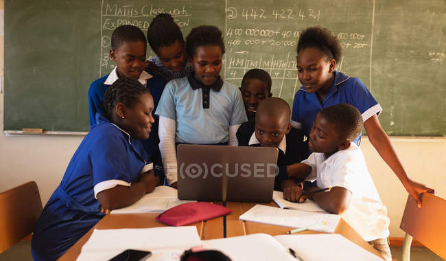 Frontansicht einer Gruppe junger afrikanischer Schüler, die vor der Tafel stehen und sitzen, die sich während einer Schulstunde in einer Township-Grundschule um einen Laptop versammelt haben — Stockfoto