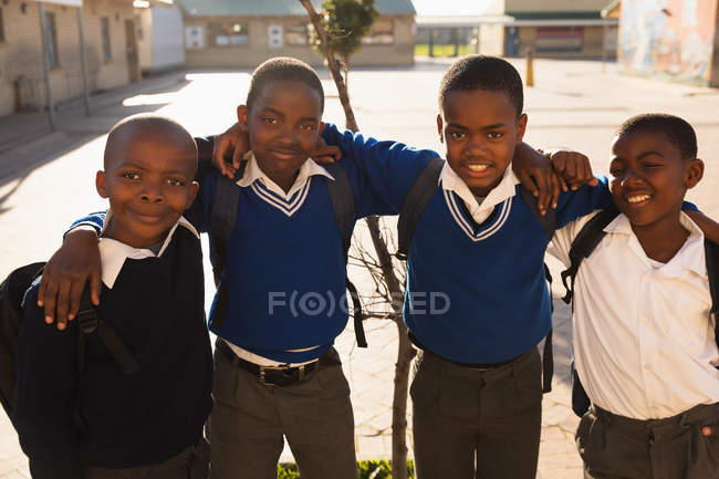 Retrato de cerca de cuatro jóvenes escolares africanos de pie con los brazos alrededor uno del otro sonriendo a la cámara en el patio de la escuela de una escuela primaria del municipio - foto de stock