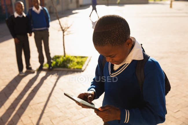 Вид сбоку на маленького африканского школьника, использующего планшетный компьютер, когда он ходит по детской площадке в школьном классе начальной школы. На заднем плане можно увидеть двух его одноклассников, наблюдающих за ним — стоковое фото