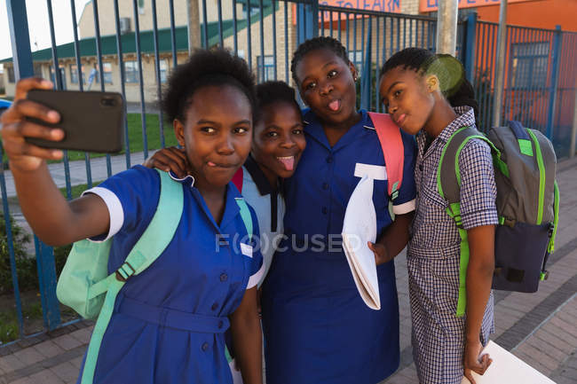 Front view close up of a group of young African schoolgirls having fun posing and taking selfies with a smartphone in a township elementary school playground — Stock Photo