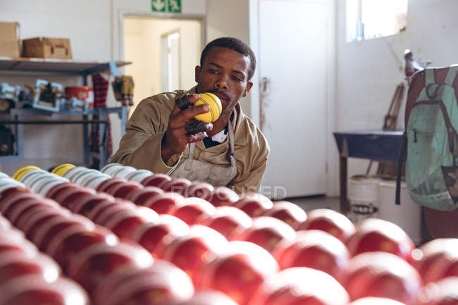 Vue de face d'un jeune Afro-Américain assis à côté de rangées de balles de cricket au bout de la chaîne de production dans une usine d'équipements sportifs. Il tient une balle de cricket jaune et l'inspecte de près. . — Photo de stock