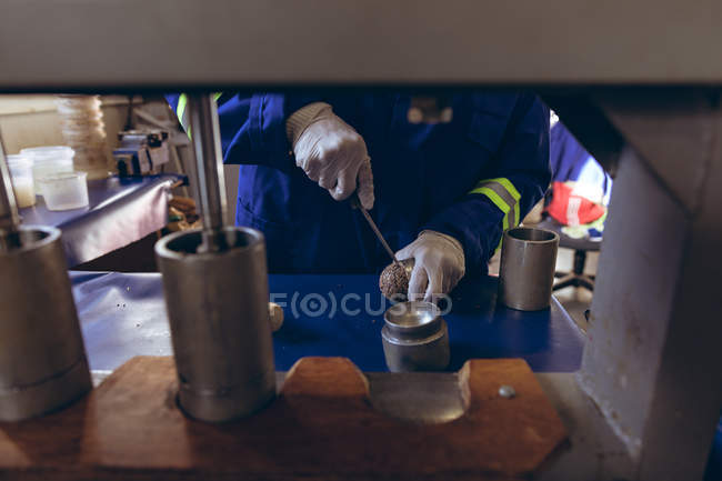 Visão frontal seção média do homem vestindo luvas e macacões segurando e cortando o núcleo de uma bola em uma fábrica fazendo bolas de críquete, visto através do equipamento . — Fotografia de Stock