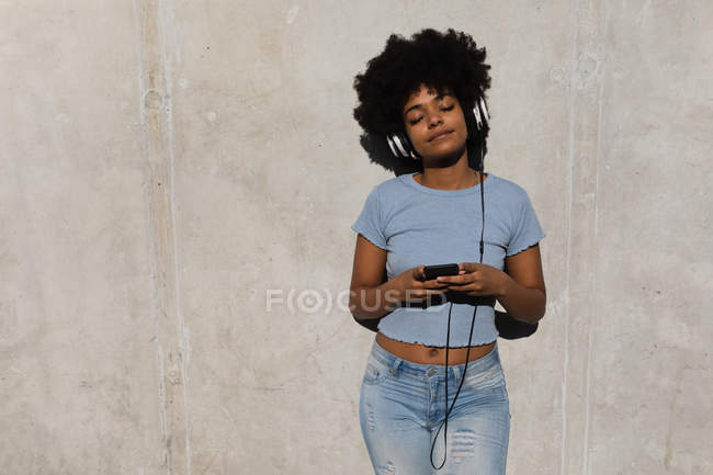 Передній вигляд молодої змішаної раси жінки, що спирається на стіну за допомогою смартфона і носить навушники, слухаючи музику на вулиці на сонці — стокове фото