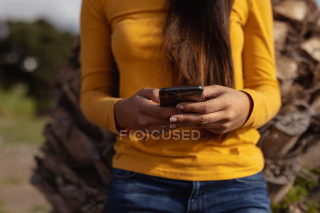 Vista frontal sección media de la mujer apoyada contra una palmera usando un teléfono inteligente - foto de stock