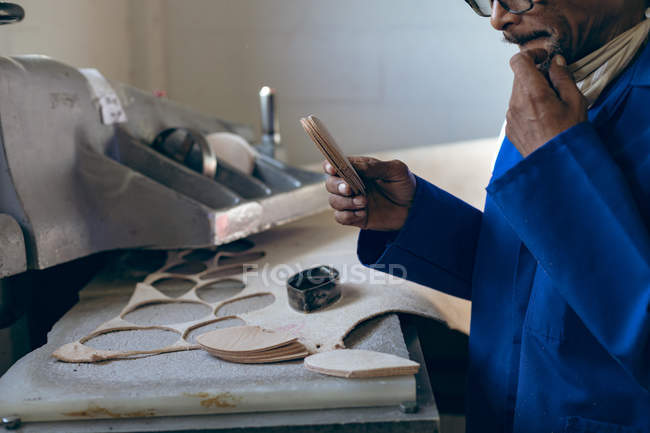 Vue latérale de la partie médiane d'un homme afro-américain d'âge moyen portant des lunettes travaillant dans une usine fabriquant des balles de cricket, regardant vers le bas et tenant des formes découpées en cuir . — Photo de stock