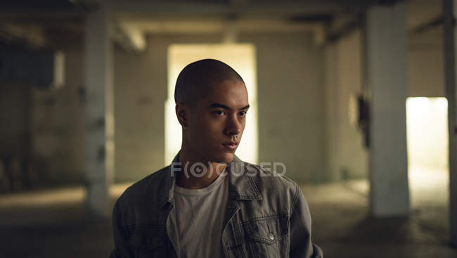 Vista frontale di un giovane ispano-americano con piercing che indossa una giacca grigia sopra una camicia bianca guardando lontano dalla telecamera all'interno di un magazzino vuoto — Foto stock
