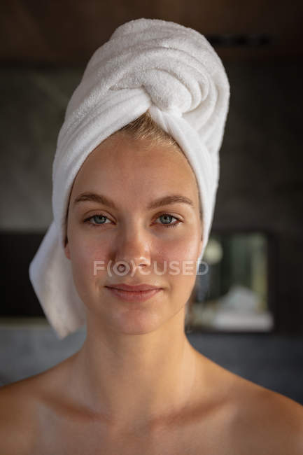 Portrait close up of a young Caucasian woman wearing a towel on her hair, looking straight to camera in a modern bathroom. — Stock Photo