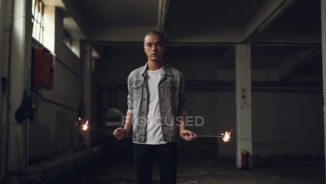 Front view of a young Hispanic-American man wearing a grey jacket over a white shirt looking intently at the camera while holding lighted sparkles on both hands inside an empty warehouse — Stock Photo