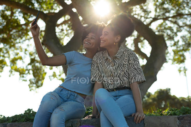 Вид спереди двух улыбающихся взрослых сестер смешанной расы, сидящих на стене в городском парке, использующих смартфон и делающих селфи с подсветкой линзами — стоковое фото