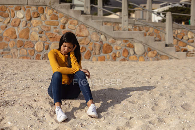 Вид спереду молода змішана жінка, що сидить на пляжі, думаючи, вирівнюючи голову на руці — стокове фото