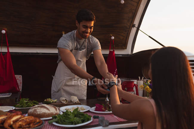 Front view close up of a young mixed race man standing serving a female customer from an open top van offering a range of takeaway food for sale, the woman is seen from the back, reaching out to take her order from him — Stock Photo