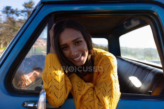 Retrato de cerca de una joven mujer de raza mixta sentada en el asiento del pasajero delantero de una camioneta, inclinada por la ventana lateral sonriendo a la cámara durante un viaje por carretera - foto de stock