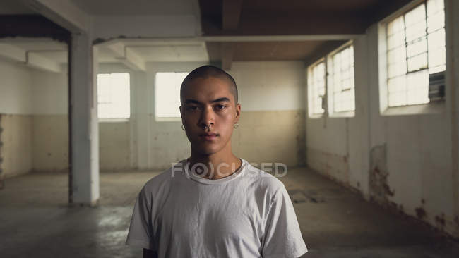 Вид спереди на молодого латиноамериканца с пирсингом в простой белой рубашке, пристально смотрящего в камеру внутри пустого склада — стоковое фото