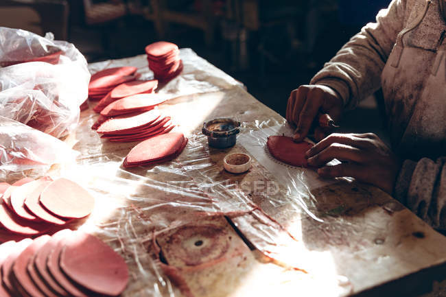 Close up of the hands of man sitting at a workbench working with cut out red leather shapes in a workshop at a factory making cricket balls — Stock Photo