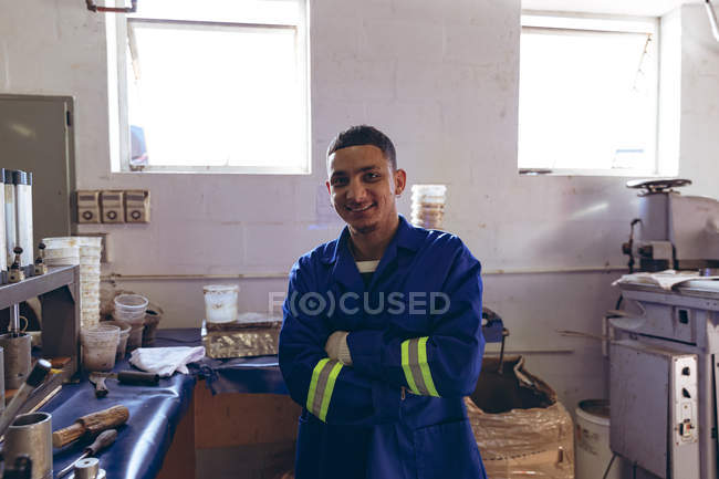 Portrait close up of a young mixed race young man wearing gloves and overalls at a factory making cricket balls, looking to camera and smiling with his arms crossed, surrounded by equipment. — Stock Photo