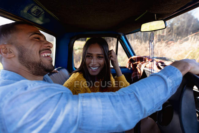 Vista lateral de una joven pareja mixta sentada en su camioneta riendo durante un viaje por carretera. El hombre está sentado al volante y la mujer lo mira y se ríe. - foto de stock