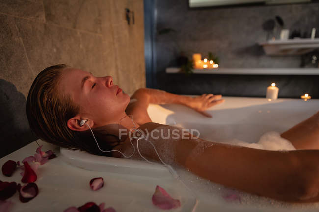 Vista lateral cercana de una joven mujer caucásica acostada en el baño con los ojos cerrados escuchando música con auriculares . - foto de stock