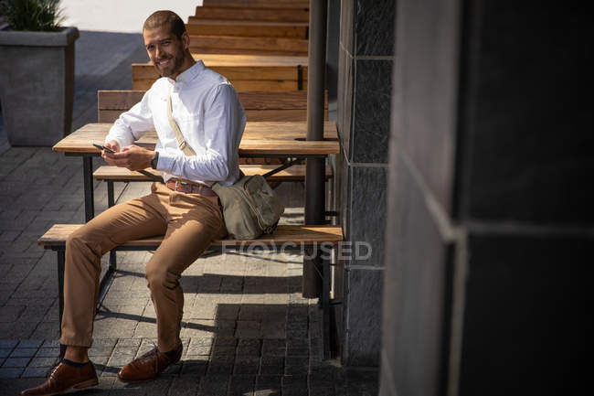 Porträt eines jungen kaukasischen Mannes, der mit seinem Smartphone eine Umhängetasche trägt und lächelnd in die Kamera blickt, auf einer Bank vor einer Bar in einer Stadtstraße. Digitaler Nomade unterwegs. — Stockfoto