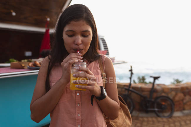 Vista frontal de cerca de una joven mestiza bebiendo una bebida de pie junto a una furgoneta que ofrece una variedad de comida para llevar en venta, la playa y el mar y una bicicleta son visibles en el fondo - foto de stock