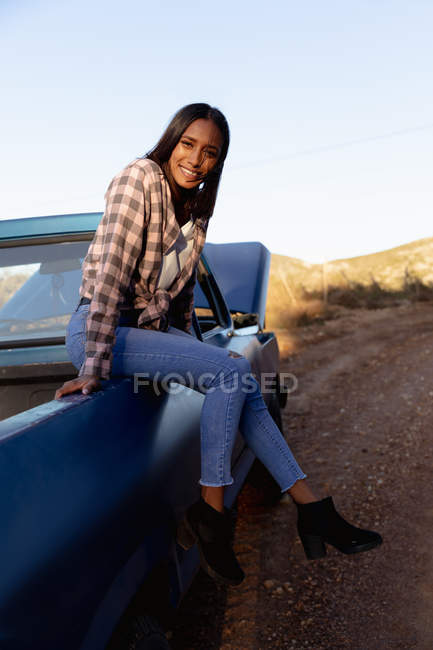 Portrait close up of a young mixed race woman sitting on the back of a pick up truck smiling to camera during a stop off on a road trip. — Stock Photo