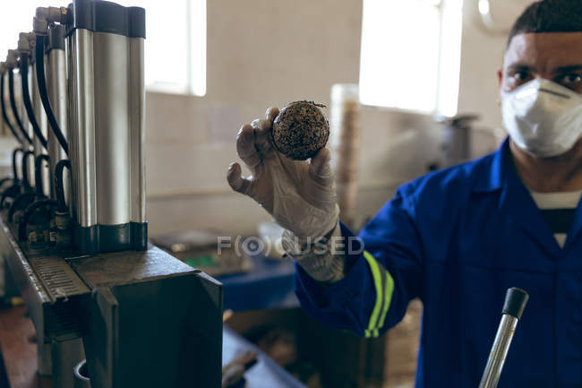 Vue de face gros plan d'un jeune homme de race mixte portant un masque facial et des gants tenant le cœur d'une balle et regardant vers la caméra dans une usine fabriquant des balles de cricket . — Photo de stock