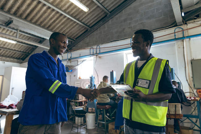 Seitenansicht von zwei afrikanisch-amerikanischen männlichen Kollegen beim Händeschütteln in einer Cricketballfabrik, einer hält einen Tablet-Computer, im Hintergrund arbeiten Menschen an Maschinen. — Stockfoto