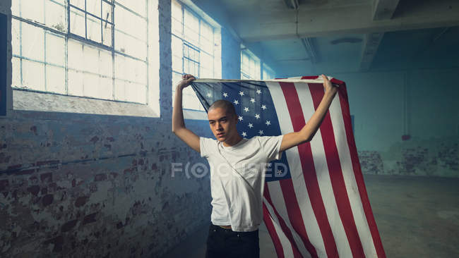 Front view of a young Hispanic-American man wearing a plain white shirt holding an American flag inside an empty warehouse — Stock Photo