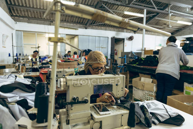 Front view of a middle aged Caucasian woman sitting and operating a sewing machine at a sports clothing factory, with colleagues visible working at sewing machines and sorting through fabrics in the background. — Stock Photo
