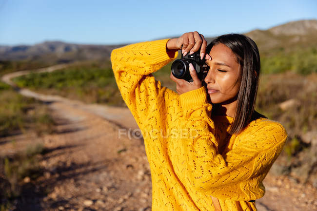 Крупный план молодой смешанной расовой женщины, фотографирующей с помощью камеры SLR на тропе в солнечном сельском пейзаже — стоковое фото