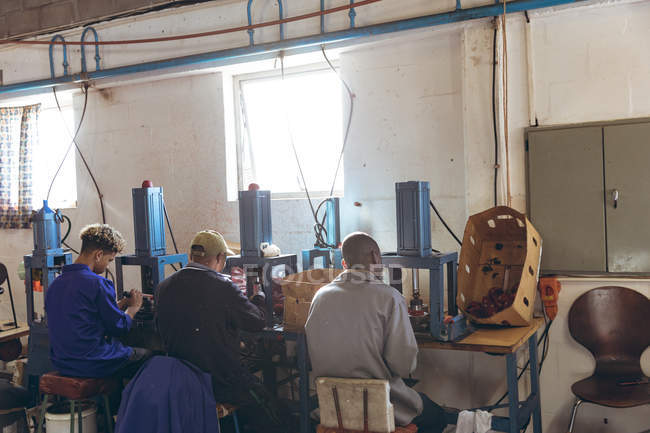 Visão traseira de um grupo diversificado de três trabalhadores do sexo masculino sentados e operando máquinas em uma fábrica de fazer bolas de críquete . — Fotografia de Stock