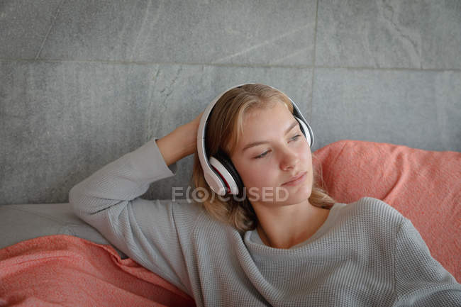 Vista frontal de cerca de una joven mujer caucásica sentada en un sofá con auriculares blancos, escuchando música y mirando hacia otro lado . - foto de stock