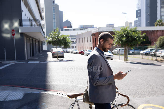 Vista lateral de un joven caucásico usando un smartphone, parado en el pavimento con su bicicleta en una calle de la ciudad. Nómada digital en movimiento . - foto de stock